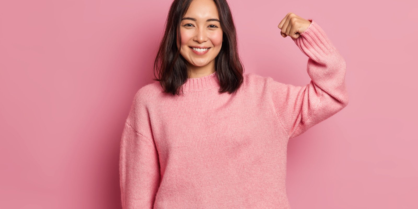 Powerful cheerful brunette woman raises arm and shows muscle demonstrates her strength looks confident at camera smiles gently wears casual long sleeved jumper isolated over pink background.